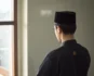 Young Muslim man in a kufi looking out a window, reflecting