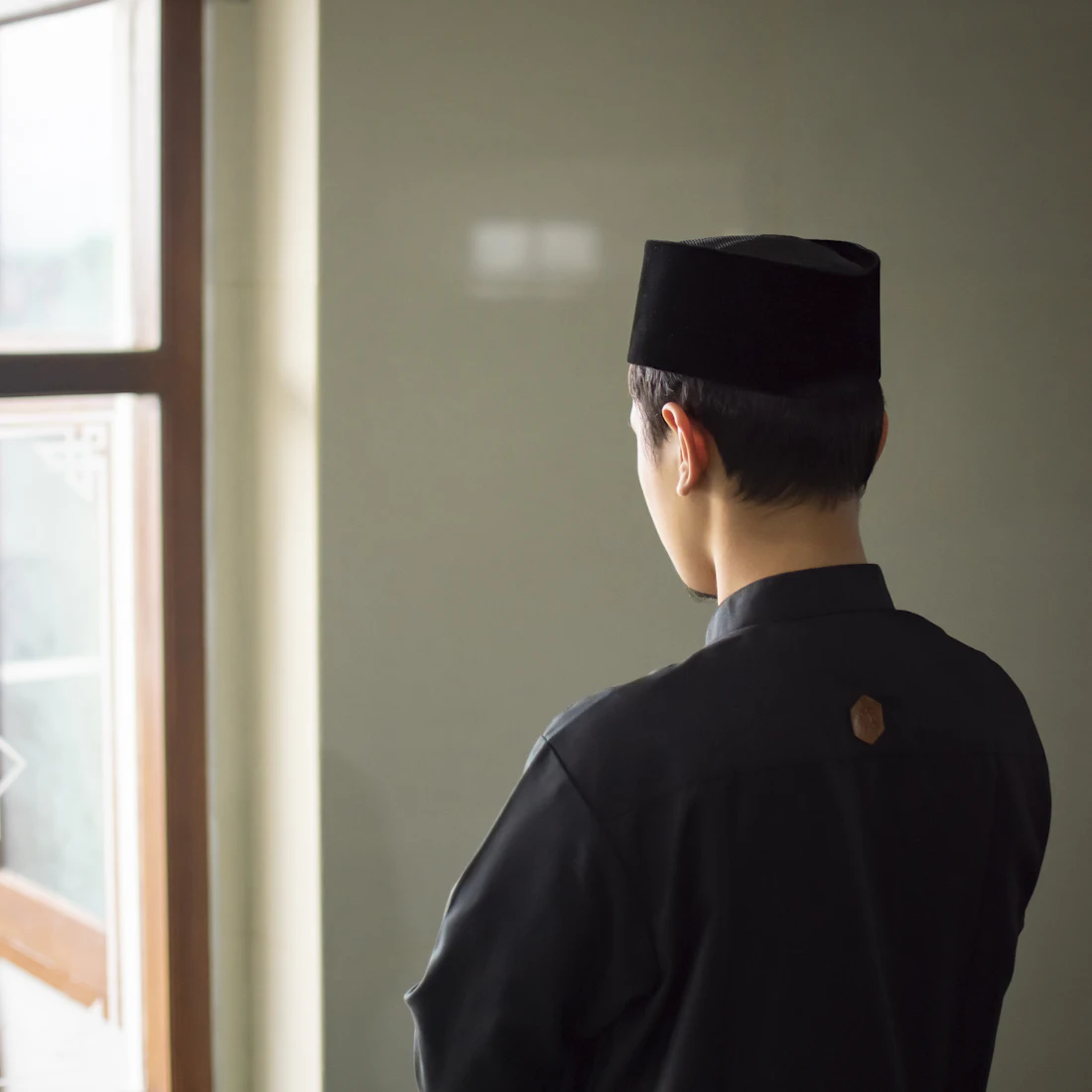 Young Muslim man in a kufi looking out a window, reflecting